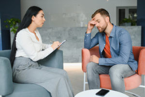 A depressed man speaks to a female therapist who is taking notes during their therapy session.