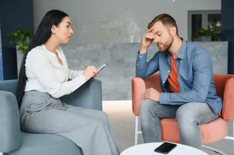 A depressed man speaks to a female therapist who is taking notes during their therapy session.