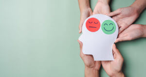 Hands holding a sad face and a happy smiley face on a brain paper cutout symbolize bipolar disorder and depression, emphasizing the mental health concept.