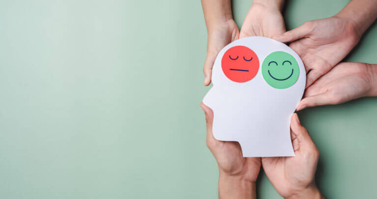 Hands holding a sad face and a happy smiley face on a brain paper cutout symbolize bipolar disorder and depression, emphasizing the mental health concept.